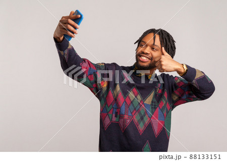 Satisfied smiling african man with dreadlocks and beard showing thumbs up looking at camera of mobile phone, video call, recommendation. Indoor studio shot isolated on gray background 88133151