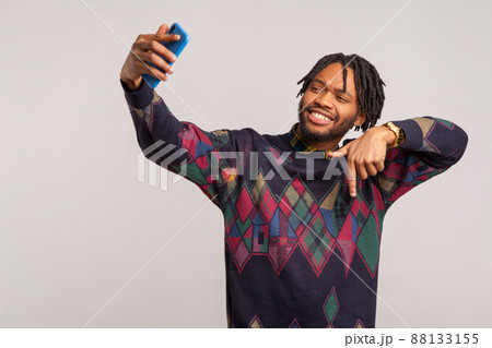 Handsome self confident african male blogger with dreadlocks showing subscribe gesture looking at camera of smartphone with toothy smile. Indoor studio shot isolated on gray background 88133155