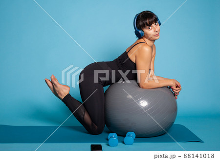 Personal trainer wearing headphones listening music while working at wellbeing stretching body muscles. Athlete fit woman in sportwear leaning on yoga fitball practicing exercices during workout Personal trainer wearing headphones listening music while working at wellbeing stretching body muscles. Athlete fit woman in sportwear leaning on yoga fitball practicing exercices during workout 88141018
