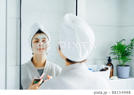 Woman in bathrobe with a towel on her head looking in the mirror and applying facial cosmetic clay mask on her face in bathroom. The concept of cosmetic procedures for skin care. Home Beauty self-care Woman in bathrobe with a towel on her head looking in the mirror and applying facial cosmetic clay mask on her face in bathroom. The concept of cosmetic procedures for skin care. Home Beauty self-care 88154998