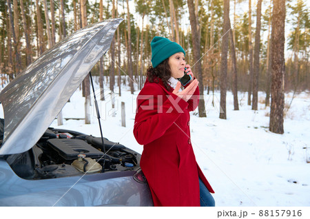 Middle aged woman in warm winter cothes sitting on the car with open hood and talks on mobile phone, calling the auto assistance while braking down in a snow covered nature during a trip by car Middle aged woman in warm winter cothes sitting on the car with open hood and talks on mobile phone, calling the auto assistance while braking down in a snow covered nature during a trip by car 88157916