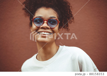 African ethnicity young woman in stylish sunglasses, with curly hair tied up in high ponytail, looking away while smiling broadly showing straight perfect teeth 88158353