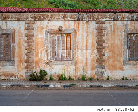 Aged house with weathered ornamental wall and shuttered windows under green hill by asphalt road 88158762