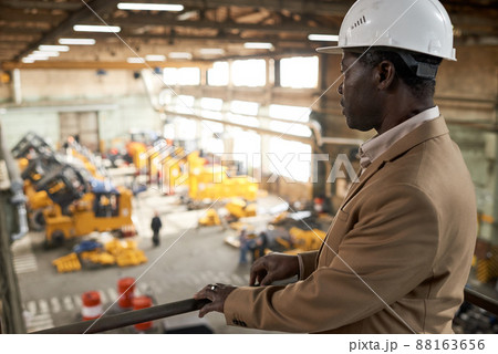 Serious African engineer in work helmet standing on balcony and watching for the work of machines in factory 88163656