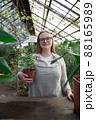 Young woman stands near table with flowers in the greenhouse and holds pot with green flower. Gardening, seedlings, sale 88165989