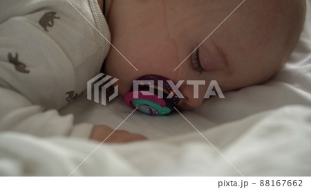 infancy, childhood, development, medicine and health concept - close-up face of newborn chubby sleeping baby 6 month with pacifier lying on stomach covered with blanket on bed on white background. infancy, childhood, development, medicine and health concept - close-up face of newborn chubby sleeping baby 6 month with pacifier lying on stomach covered with blanket on bed on white background. 88167662