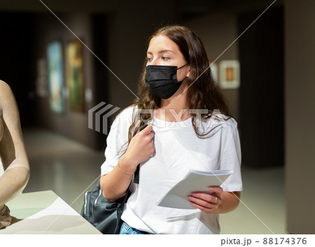 Portrait of a young girl visitor in a protective mask, looking at a sculpture in the museum hall 88174376