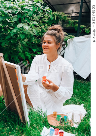 Artist painting on the easel outdoors in the garden. Open air outdoor art workshop. Draw on the canvas with brush and palette sitting on the grass during a picnic in a park 88174905