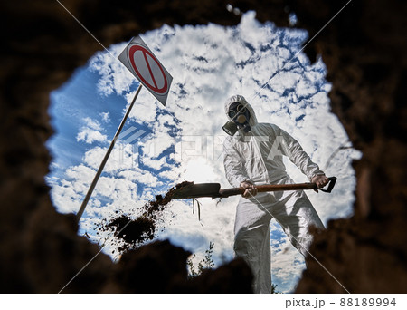 View inside pit of scientist in white suit and gas mask standing near prohibition sign and digging ground to obtain samples and study ground cover in contaminated area. Cloudy sky on the background. 88189994