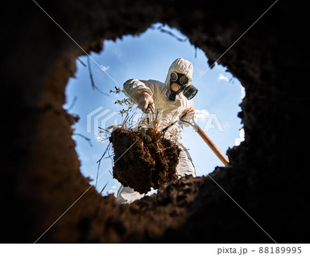View from inside pit of ecologist digging pit by shovel and planting tree, wearing gas mask and protective overalls outdoors, on background of blue cloudy sky. View from inside pit of ecologist digging pit by shovel and planting tree, wearing gas mask and protective overalls outdoors, on background of blue cloudy sky. 88189995