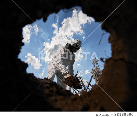 View from inside pit of ecologist digging pit by shovel and planting tree, wearing gas mask and protective overalls outdoors, on background of blue cloudy sky. View from inside pit of ecologist digging pit by shovel and planting tree, wearing gas mask and protective overalls outdoors, on background of blue cloudy sky. 88189996