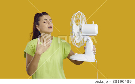 Exhausted young lady having headache because of summer heat, holding fan in her hand is exhausted from the heat, touching her forehead, yellow studio background, need for air conditioner 88190489