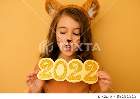 A girl with ears and tiger whiskers painted on face looks down in surprise at the numbers of 2022. The enthusiastic expression on the female face. In anticipation of the New Year A girl with ears and tiger whiskers painted on face looks down in surprise at the numbers of 2022. The enthusiastic expression on the female face. In anticipation of the New Year 88191515