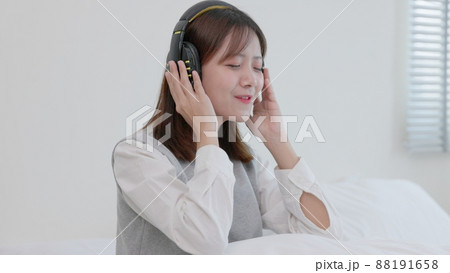 While sitting on her bed at home, a happy young Asian woman in earphones sings and listens to music. Girl dances while listening to music through headphones. A content woman spends her evenings in While sitting on her bed at home, a happy young Asian woman in earphones sings and listens to music. Girl dances while listening to music through headphones. A content woman spends her evenings in 88191658