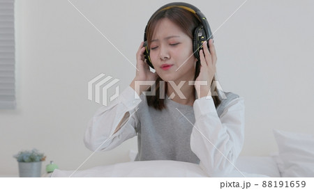 While sitting on her bed at home, a happy young Asian woman in earphones sings and listens to music. Girl dances while listening to music through headphones. A content woman spends her evenings in While sitting on her bed at home, a happy young Asian woman in earphones sings and listens to music. Girl dances while listening to music through headphones. A content woman spends her evenings in 88191659