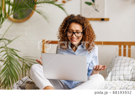 Joyful emotional african american teenage girl feeling excited while sitting on bed with laptop during video call Joyful emotional african american teenage girl feeling excited while sitting on bed with laptop during video call 88193932