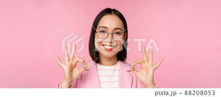 Close up portrait of impressed corporate woman, asian business lady in glasses, showing okay sign, looking amazed at camera, recommending, pink background 88208053