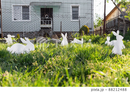 The concept of Easter holidays. Decorative Handicrafts handmade in the form of white rabbits and colorful eggs lying on the grass. House in the background. Copy space 88212488