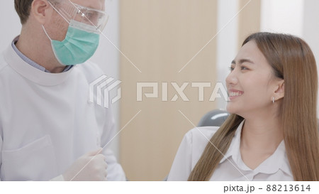 At a sophisticated dental facility, a Caucasian male dentist with a face mask explains how to treat teeth to a cheerful Asian woman patient. 88213614