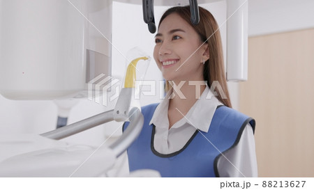 On a panoramic x-ray machine, the dentist aligns the bite of a woman. In a dental clinic, a woman dressed in a lead apron stands still as a panoramic X-ray machine circles her head.. On a panoramic x-ray machine, the dentist aligns the bite of a woman. In a dental clinic, a woman dressed in a lead apron stands still as a panoramic X-ray machine circles her head.. 88213627