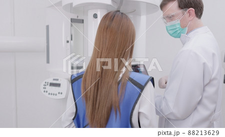 On a panoramic x-ray machine, the dentist aligns the bite of a woman. In a dental clinic, a woman dressed in a lead apron stands still as a panoramic X-ray machine circles her head.. On a panoramic x-ray machine, the dentist aligns the bite of a woman. In a dental clinic, a woman dressed in a lead apron stands still as a panoramic X-ray machine circles her head.. 88213629