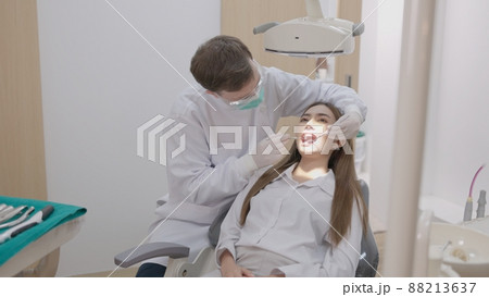 In a dental clinic, a side view of a dentist in uniform treating the teeth of a female patient with a mirror. A dentist works on a gorgeous woman's teeth while holding a dental mirror. Dentistry as a In a dental clinic, a side view of a dentist in uniform treating the teeth of a female patient with a mirror. A dentist works on a gorgeous woman's teeth while holding a dental mirror. Dentistry as a 88213637