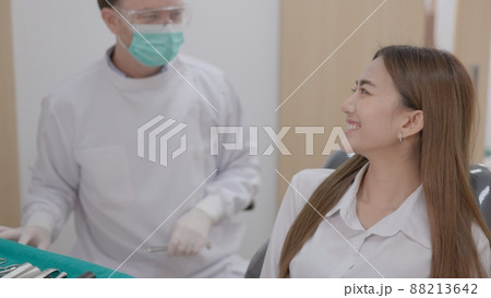 At a sophisticated dental facility, a Caucasian male dentist with a face mask explains how to treat teeth to a cheerful Asian woman patient. At a sophisticated dental facility, a Caucasian male dentist with a face mask explains how to treat teeth to a cheerful Asian woman patient. 88213642
