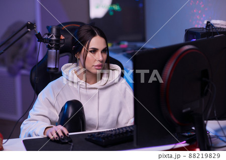 Happy woman putting on headphones and playing computer game. Female gamer in glasses sitting and looking at computer monitor. Happy woman putting on headphones and playing computer game. Female gamer in glasses sitting and looking at computer monitor. 88219829