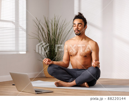 Relaxed black man meditating in lotus pose in front of laptop at home Relaxed black man meditating in lotus pose in front of laptop at home 88220144