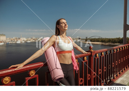 Happy peaceful woman with mat and backpack leans onto handrail on city embankment Happy peaceful woman with mat and backpack leans onto handrail on city embankment 88221579
