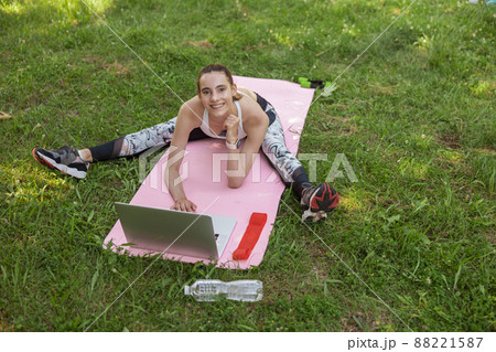 Attractive athletic lady in tracksuit does stretching exercise near open laptop on grass 88221587