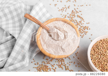 Buckwheat flour in a wooden bowl with wooden spoon and raw green buckwheat grain on grey stone background, top view 88226080