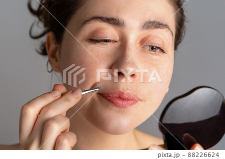 A young woman looks in the mirror and plucks her hair over her upper lip with tweezers. The concept of getting rid of unwanted facial hair. Close up 88226624