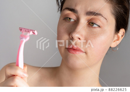 A young pretty woman with a dissatisfied expression is holding a razor in her hands.The concept of getting rid of unwanted hair. 88226625
