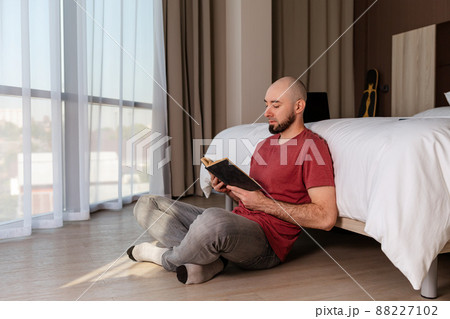 A young, bearded, bald man sits cross-legged on the floor of his house, reading a book. Indoor. The concept of education and reading 88227102