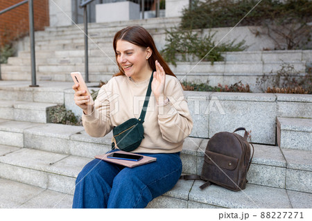 Beautiful smiling female student making a selfie with smartphone outdoors. Phone Communication. Happy cheerful young woman walking on city street, Urban lifestyle concept. Traveler 88227271