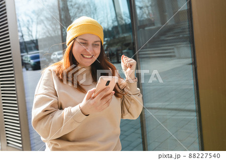 Portrait of a satisfied young woman holding mobile phone and celebrating outdoors. Happy girl walking on city street, hipster female in yellow hat overjoyed with winning online contest on web site Portrait of a satisfied young woman holding mobile phone and celebrating outdoors. Happy girl walking on city street, hipster female in yellow hat overjoyed with winning online contest on web site 88227540
