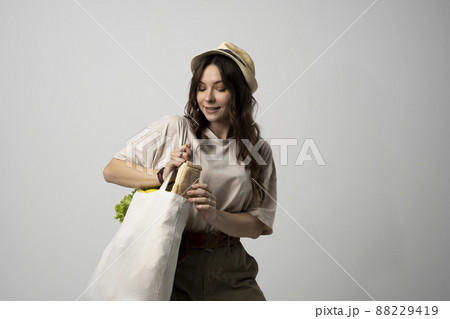 Smiling young woman in beige t-shirt and a hst with a mesh eco bag full of vegetables and on a white studio background. Sustainable lifestyle. Eco friendly concept. Zero waste. 88229419