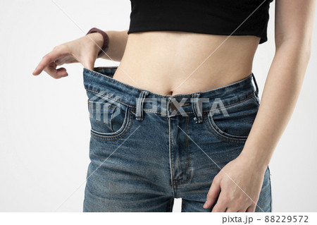 Woman in blue oversize jeans showing a result of weight loss on white background. Diet concept and weight loss. Woman in blue oversize jeans showing a result of weight loss on white background. Diet concept and weight loss. 88229572