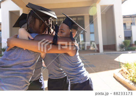 Multiracial elementary schoolboys wearing mortarboard while making huddle against school building Multiracial elementary schoolboys wearing mortarboard while making huddle against school building 88233173