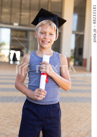 Portrait of happy caucasian elementary schoolboy with mortarboard and degree standing against school Portrait of happy caucasian elementary schoolboy with mortarboard and degree standing against school 88233181