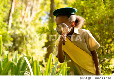 African american scout girl in uniform looking at plants through magnifying glass in forest 88233748