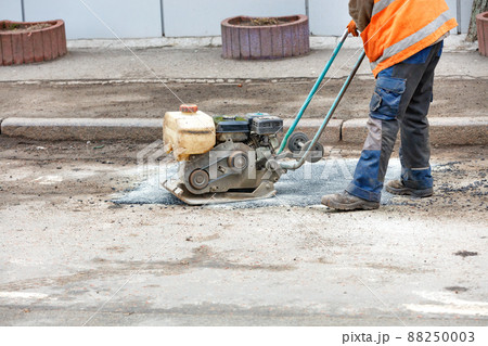 A road worker is patching up a pothole in an old road using an old petrol plate compactor. 88250003