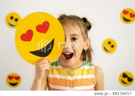 A little girl, smiling with her mouth open, holds in her hands a cardboard love smile with hearts instead of eyes, covering part of her face with them. World emoji day. Anthropomorphic smile Face. A little girl, smiling with her mouth open, holds in her hands a cardboard love smile with hearts instead of eyes, covering part of her face with them. World emoji day. Anthropomorphic smile Face. 88256657