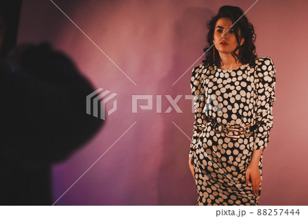 Beautiful brunette fashion model in polka dot dress posing for the photographer on a pink background. Shadow and glare on the wall 88257444