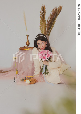 Beautiful brunette girl in a white jacket and white tights in a light interior holding a rose bouquet 88257470