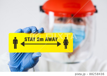 Medical worker in personal protective equipment holding yellow "STAY 2m AWAY" sign 88258436