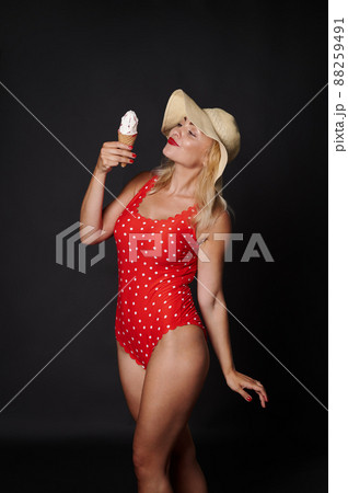 Sexy stunning blonde woman in summer straw hat and red swimsuit provocatively eating a delicious ice-cream cone with strawberry flavour, isolated on black background. Summer concept with copy space Sexy stunning blonde woman in summer straw hat and red swimsuit provocatively eating a delicious ice-cream cone with strawberry flavour, isolated on black background. Summer concept with copy space 88259491