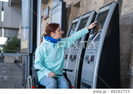 Caucasian woman in a wheelchair buys a train ticket at a self-service checkout. Caucasian woman in a wheelchair buys a train ticket at a self-service checkout. 88262755