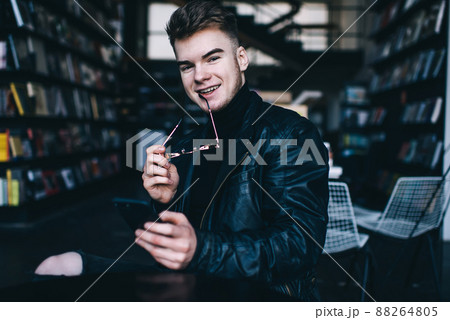 Positive young man with smartphone looking at camera in library Positive young man with smartphone looking at camera in library 88264805